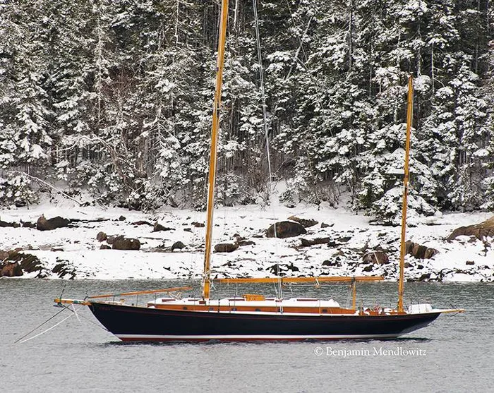 Glide afloat, a black-hulled ketch lying off a snow-covered Maine shore.
