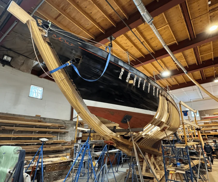 The hull from astern in the shop, shored on stands with new framing visible inside.